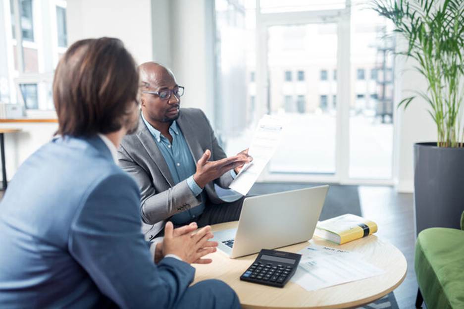 In a business meeting, two professionals review paperwork over a laptop, calculator, and book. The meeting is the main content of the page.